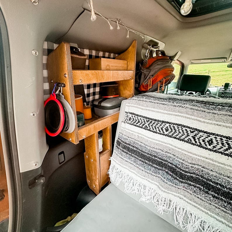 May include: A wooden shelf unit with two shelves is mounted in the back of a vehicle. The shelves are filled with various items, including a black and gray case, a brown box, and a few other items. A black and white blanket is draped over the back seat of the vehicle.