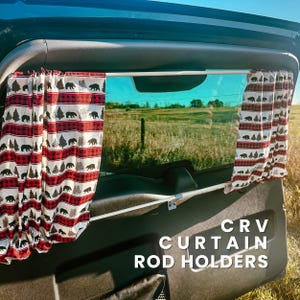Può includere: Un finestrino di un veicolo con un'asta per tende e tende. Le tende presentano un motivo a quadri rossi e neri con sagome di orsi e alberi neri. Il testo "CRV CURTAIN ROD HOLDERS" è visibile sulla finestra.