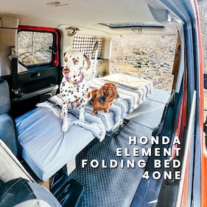 Puede incluir: Un dálmata y un perro salchicha están sentados en una cama plegable dentro de un Honda Element. La cama está cubierta con una manta azul y una manta estampada. El texto "HONDA ELEMENT FOLDING BED 4 ONE" es visible.