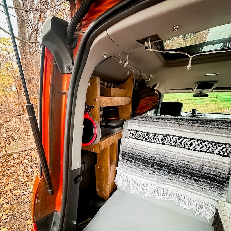 May include: The interior of a car with a wooden shelf system and a black and white striped blanket on the back seat. The shelf system has a red bucket hanging from it.