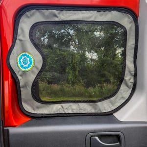 May include: A grey mesh vehicle window screen with a black border installed on a red car door. The screen features a circular fabric patch with a blue, green, and white floral pattern on the left side, designed to fit the window frame.
