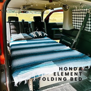 Peut inclure: Un Honda Element avec un lit pliant à l'arrière. Le lit est fait avec une couverture rayée bleu et blanc et des oreillers. Le texte "HONDA ELEMENT FOLDING BED" est visible sur l'image.