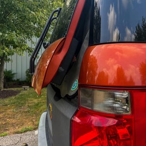 May include: A metallic orange vehicle rear section featuring a partially open tailgate, a black ladder attached to the side, a red and clear taillight assembly, and a black trailer hitch ball mount extending from the lower bumper area.