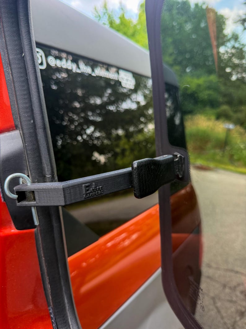 May include: A black 3D-printed plastic vent latch for a vehicle window, featuring the text "ELITE CAMPERS" embossed on the arm. The mechanism includes a metal pin and a textured handle, attached to the frame of an orange vehicle window.