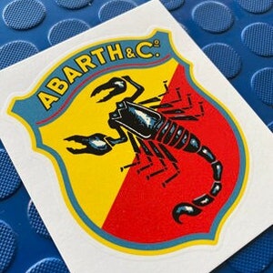 May include: A yellow and red shield with a blue outline and a black scorpion with a raised tail. The shield has the text "ABARTH & C." in blue.