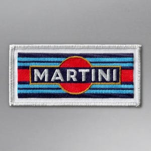 Peut inclure: Écusson brodé rectangulaire avec le mot "MARTINI" en lettres dorées. Le design comprend un cercle rouge et des rayures bleues et rouges. L'écusson a une bordure blanche.
