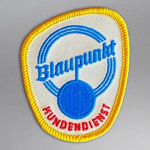 Pode incluir: Patch bordado com fundo branco e borda amarela. O patch apresenta a palavra "Blaupunkt" em azul, arqueada sobre um círculo azul. Abaixo, a palavra "HUNDENDIENST" está em vermelho.