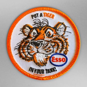 May include: A white embroidered patch with an orange border. The patch features a cartoon tiger with the text "Put a Tiger in Your Tank!" and the Esso logo.