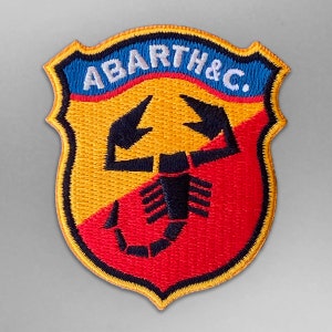 May include: Embroidered patch with the text "ABARTH & C." in white on a blue background. The patch features a shield design with a yellow and red background and a black scorpion emblem.