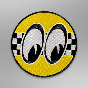 May include: A yellow and black embroidered patch with two white eyes with black pupils. The eyes are set against a yellow background with a black and white checkered border.