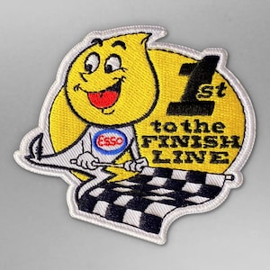 Può includere: Un personaggio dei cartoni animati giallo che tiene una bandiera a scacchi con il testo "1° al traguardo" e il logo Esso. Il personaggio indossa un cerchio blu e bianco con la parola "Esso" al suo interno.
