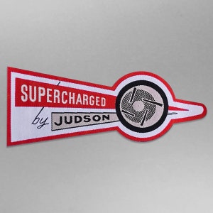 Peut inclure: Écusson brodé rouge et blanc avec le texte "SUPERCHARGED by JUDSON" et une illustration en noir et blanc d'une pièce de moteur stylisée.