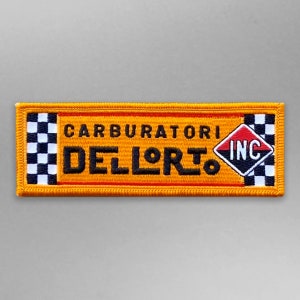 May include: A yellow and black embroidered patch with the text "CARBURATORI DELL'ORTO INC" in black lettering. The patch has a checkered pattern on the left and right sides.