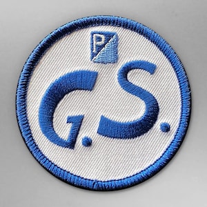 Puede incluir: Un parche circular blanco con un borde azul. El parche tiene un "G.S." bordado en azul con un pequeño cuadrado azul con una "P" en la esquina superior izquierda.