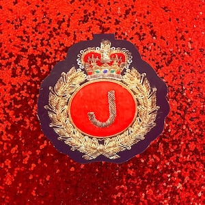 May include: A circular patch featuring a dark blue background with a gold embroidered laurel wreath surrounding a bright red center circle. The letter J is embroidered in gold in the middle, topped by a detailed gold crown with small colored accents.