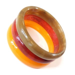 Puede incluir: Tres pulseras apiladas en tonos ámbar, rojo y marrón. Las pulseras son lisas y brillantes, con forma redondeada. Los colores van de un naranja brillante a un rojo intenso y luego a un marrón veteado.