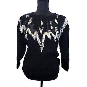May include: Black sweater with long sleeves. The sweater features a decorative design across the chest and shoulders, with a mix of silver and white patterned fabric, black beads, and black mesh. The sweater is displayed on a black mannequin.