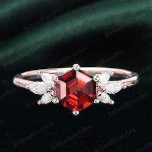 May include: A rose gold engagement ring featuring a hexagonal red gemstone surrounded by smaller white gemstones. The ring is displayed on a green fabric background.