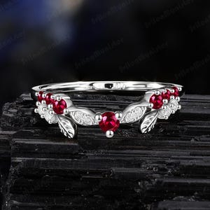 Peut inclure: Une bague en argent avec un motif floral. La bague est ornée de pierres précieuses rouges et de petites pierres précieuses blanches. La bague est posée sur un fond sombre.