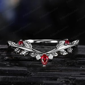 Könnte beinhalten: Ein silberner Ring mit Blattmuster, besetzt mit drei roten Edelsteinen und zwei kleinen weißen Steinen. Der Ring hat ein gebogenes Band. Der Hintergrund ist dunkel und strukturiert.