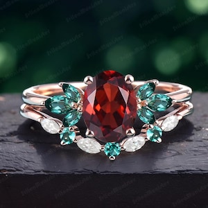 May include: A rose gold engagement ring set with a large oval red gemstone surrounded by smaller green gemstones and white diamonds. The ring is on a dark surface.