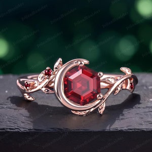 May include: A rose gold ring featuring a large, hexagonal, red gemstone set within a delicate, leafy design. The ring incorporates a crescent moon shape and smaller red gemstones on the leaves.