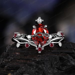 Peut inclure: Bague en argent ornée d'une grosse pierre rouge en forme de poire. La bague présente de plus petites pierres précieuses rouges et blanches, et un motif décoratif en forme de vigne. La bague est posée sur un fond sombre.