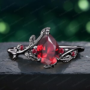 Peut inclure: Une bague noire avec une pierre rouge en forme de poire entourée de petites pierres rouges. La bague présente un délicat motif de feuilles.