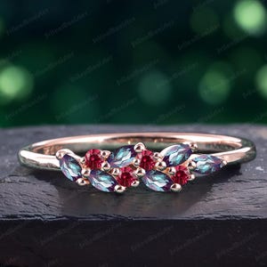 May include: A delicate rose gold band ring featuring a cluster of marquise-shaped gemstones. The ring incorporates a mix of deep red and blue-green gemstones, creating a vibrant and eye-catching design.