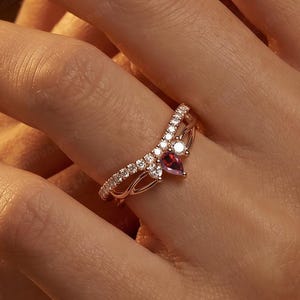Puede incluir: Un anillo de oro rosa con una banda en forma de V adornada con pequeñas piedras claras y una piedra preciosa central en forma de pera de color rosa rojizo. La banda metálica tiene un diseño calado intrincado que se curva hacia arriba.
