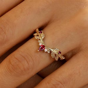 Könnte beinhalten: Ein goldener V-förmiger Ring mit einem zarten Blattmotiv entlang der Ringschiene. Das Design ist mit einem zentralen birnenförmigen roten Edelstein und zwei kleineren runden roten Edelsteinen akzentuiert, die zwischen kleinen klaren Steinen liegen.