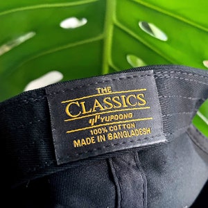 May include: A black baseball cap with a yellow label that reads "THE CLASSICS YUPOONG 100% COTTON MADE IN BANGLADESH"