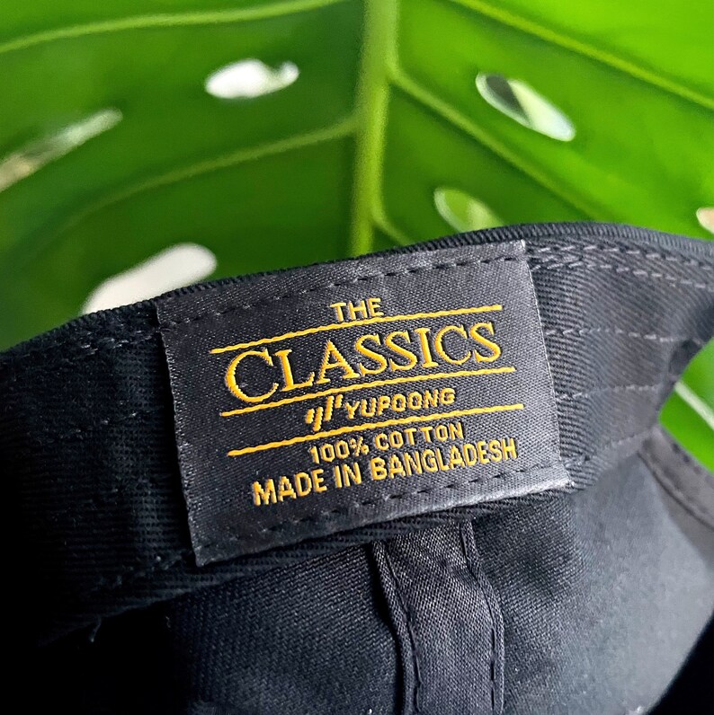 May include: A black baseball cap with a yellow label that reads "THE CLASSICS YUPOONG 100% COTTON MADE IN BANGLADESH"