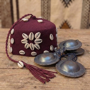 May include: A burgundy fez hat decorated with white seashells and a matching tassel. Beside the hat are four silver-colored finger cymbals. The items are arranged on a wooden surface.