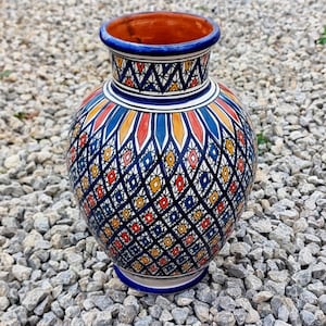 Op de afbeelding: Een handbeschilderde keramische vaas met een bolvormig lichaam en een uitlopende rand. De vaas heeft een levendig geometrisch patroon in blauw, rood, geel en oranje op een donkerblauwe achtergrond. De binnenkant is warm oranje.