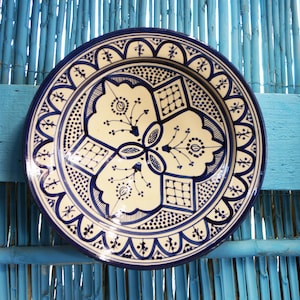 May include: A round, decorative ceramic plate with a cream-colored center and intricate blue patterns. The design features floral and geometric motifs, including dots, lines, and stylized flowers. The plate has a blue rim and is displayed against a blue background.