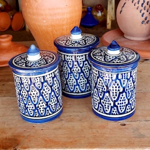Puede incluir: Tres tarros cilíndricos de cerámica con tapa, que presentan una base blanca y complejos patrones azules pintados a mano de arcos y puntos repetidos. Cada tarro tiene una tapa a juego con un pomo puntiagudo de color azul y blanco.