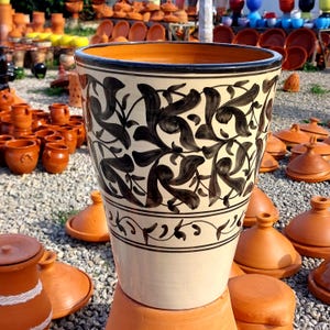 Puede incluir: Un macetero de cerámica blanca con un diseño floral negro. El macetero tiene una base ancha y una parte superior estrecha, y está decorado con una banda negra alrededor del borde.