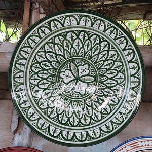 以下が含まれることがあります： 白と緑の花柄模様の大きな丸いセラミックプレート。プレートは濃い緑色の縁と中央に複雑な模様が特徴です。デザインには様式化された花と葉のような形が含まれており、装飾的で芸術的な外観を作り出しています。