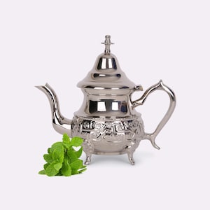 Puede incluir: Una tetera plateada con detalles ornamentados y una boquilla, asa y patas curvas. Hojas de menta verde fresca se colocan junto a la tetera. La tetera tiene una tapa decorativa y un diseño clásico.