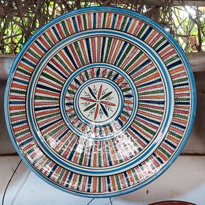 Może przedstawiać: Duży, okrągły, ręcznie malowany ceramiczny talerz o żywym, kolorowym wzorze. Talerz ma jasnoniebieską obwódkę i centralny wzór z promienistymi paskami w kolorze pomarańczowym, zielonym, niebieskim i czerwonym. Środek ma wzór gwiazdy.