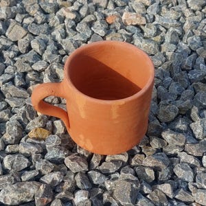 Peut inclure: Une tasse en terre cuite avec une anse. La tasse est posée sur un lit de petites pierres grises.