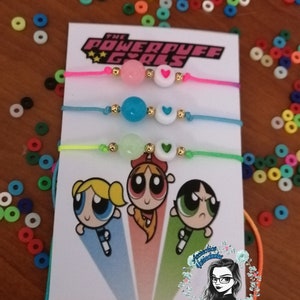 Puede incluir: Tres pulseras de amistad coloridas con cuentas y detalles de corazón se exhiben en una tarjeta con el logotipo y los personajes de las Supernenas. Las pulseras son rosas, azules y verdes, con detalles dorados.