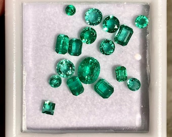 May include: A collection of vibrant green emerald gemstones in various shapes, including round, oval, and emerald cuts. The gemstones are displayed in a white square container, showcasing their clarity and brilliance. The emeralds vary in size.