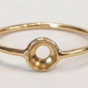 May include: A gold ring with a circular setting designed to hold a gemstone. The ring has a thin band and a round setting with a central hole. The ring is made of shiny gold metal.