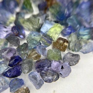 May include: A collection of raw, uncut gemstones in various shades of blue, green, and purple. The stones have a rough, natural texture and are scattered across a white surface. The gemstones vary in size and shape, with some appearing translucent.