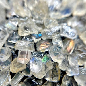 May include: Close-up of a pile of small, clear, rough-cut gemstones. The stones have a crystalline appearance with subtle color variations, including hints of blue, green, and gold. The gemstones are scattered, creating a textured surface.