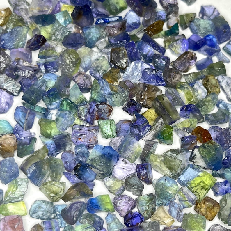 May include: A collection of raw, uncut gemstones in various shades of blue, green, and purple. The stones are irregularly shaped and appear to be scattered across a white surface. The gemstones vary in size and have a translucent quality.