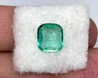 May include: A square-cut, vivid green emerald gemstone. The gem is set on a white, textured fabric. The emerald has a polished surface and a slight translucence, showcasing its natural colour and clarity. The gemstone is held by a person.