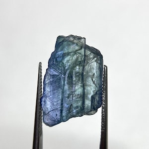 May include: A raw, translucent kyanite crystal displaying a gradient of deep blue to pale blue and green hues. The stone features a jagged, elongated shape with visible vertical striations and internal fractures throughout its crystalline structure.